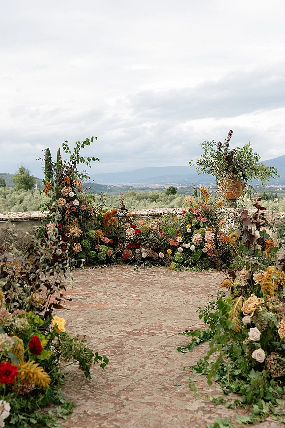 Ceremony aisle decor with a floral aisle runner of roses, dahlias, and greenery garlands along a stone terrace with hills beyond
