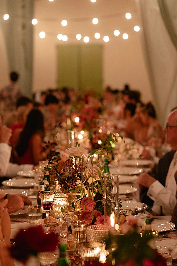 Reception tablescape on a long banquet table with greenery garland, floral centerpiece, taper candles, and string lights under white tent draping