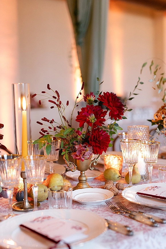 Reception tablescape with a wedding table centerpiece of red dahlias in a brass compote, taper candles, crystal goblets, pears under warm tent lighting