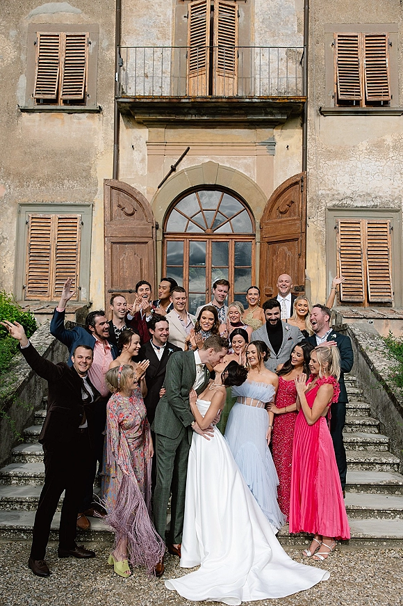 Wedding party photo of bride and groom kissing as friends cheer on stone steps by an arched doorway at a historic building facade
