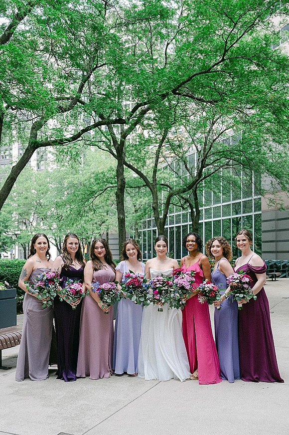 Bridesmaid group photo with bride with bridesmaids holding bouquets in bright dresses, posing on a city sidewalk by glass building and trees