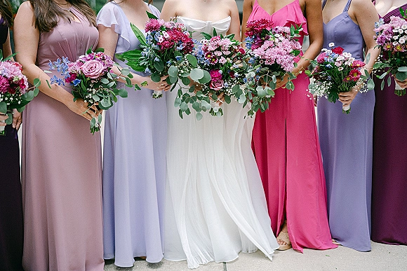 Bridesmaid bouquets of bright pink and purple flowers with rose and eucalyptus accents held beside a bride in white on outdoor pavement