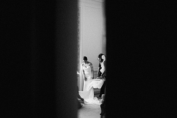 Bridal getting ready as bridesmaids help bride getting into dress, adjusting the wedding dress train by window light in a molded room