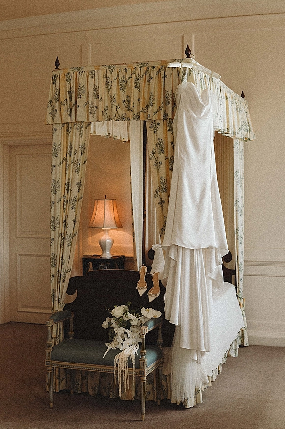 Wedding dress hanging with veil over a four poster bed, bridal shoes and bouquet arranged below in a classic paneled bedroom