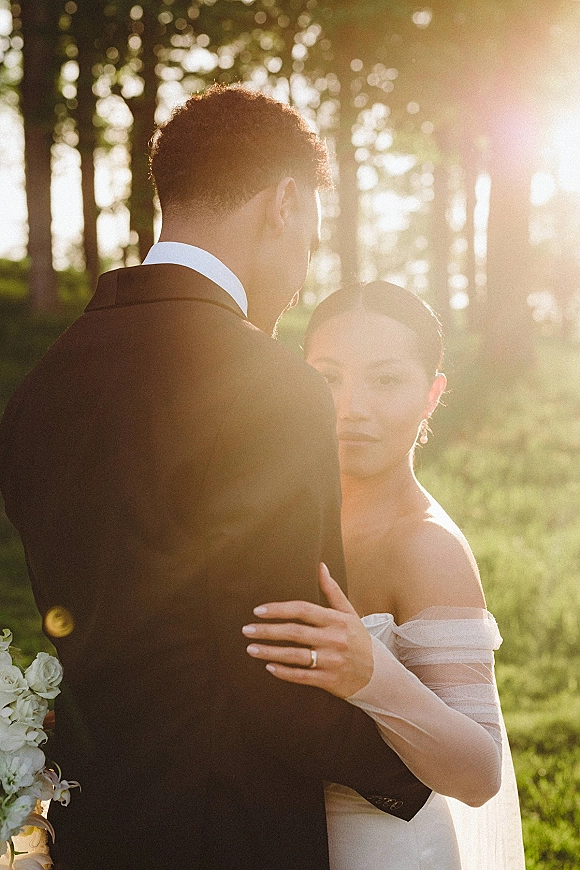 Couple portrait of bride and groom embrace, bride looking over shoulder with veil and bouquet, backlit by golden sun in trees