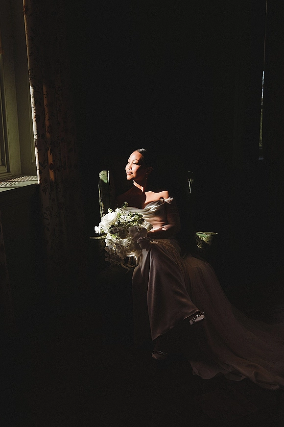 Bridal portrait of a bride sitting in chair by window light, wearing a strapless wedding dress and gloves, holding a white bouquet in a dark room