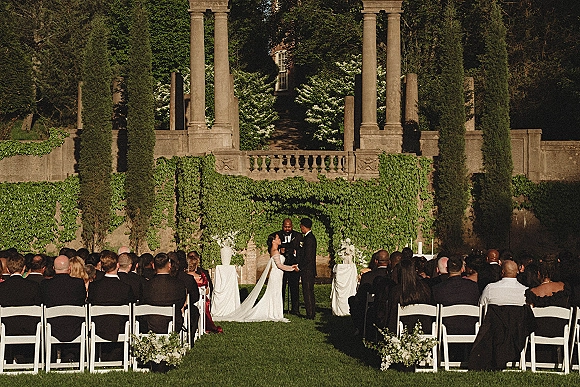 Wedding ceremony with bride and groom exchanging vows before an officiant, framed by an ivy-covered stone wall and white chairs