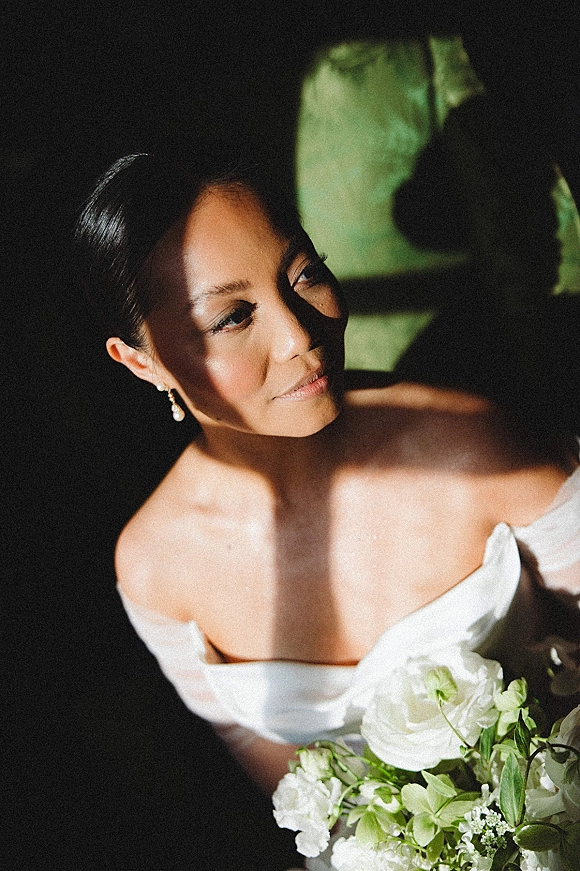 Bridal portrait of a bride looking up in an off the shoulder wedding dress, holding a white and greenery bouquet in window light indoors