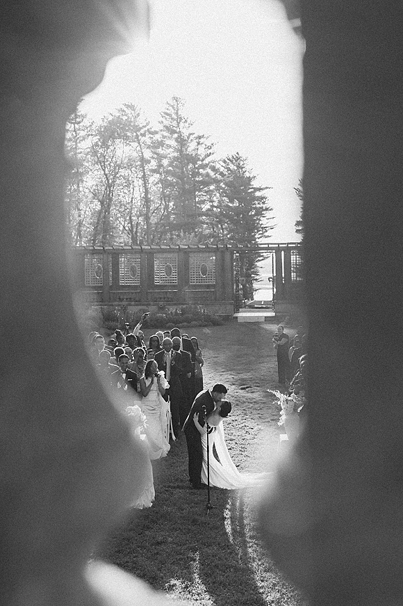 Wedding kiss during an outdoor wedding ceremony as the bride in veil holds a bouquet and guests watch on a sunlit lawn under trees