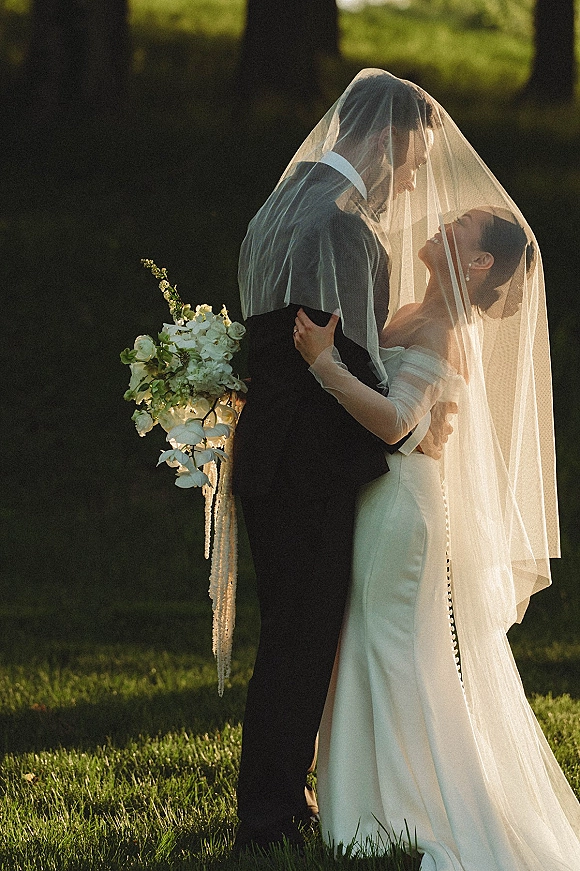 Wedding couple portrait of bride and groom under veil, embracing on a sunlit lawn with trees, bride holding a cascading white bouquet