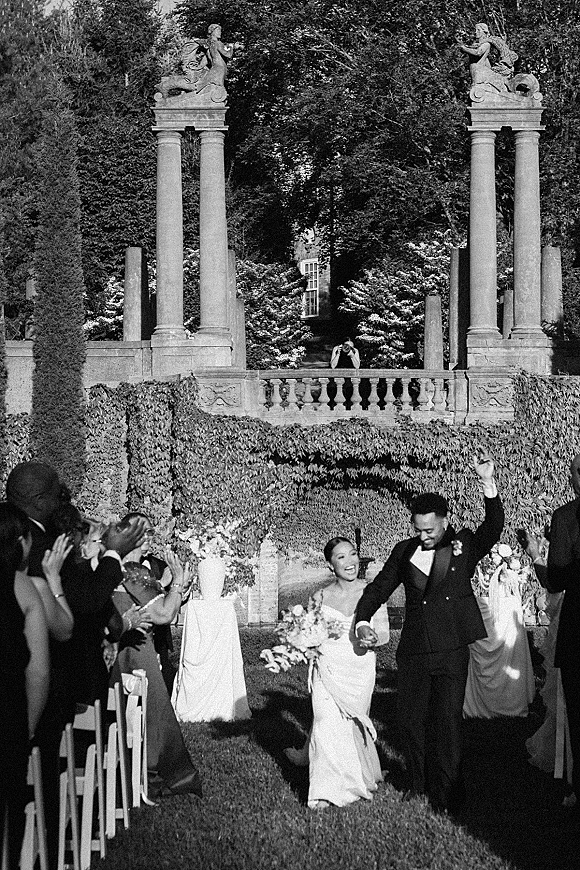 Wedding recessional as bride and groom hold hands walking the aisle, bouquet and tuxedo, guests cheering in a columned garden setting