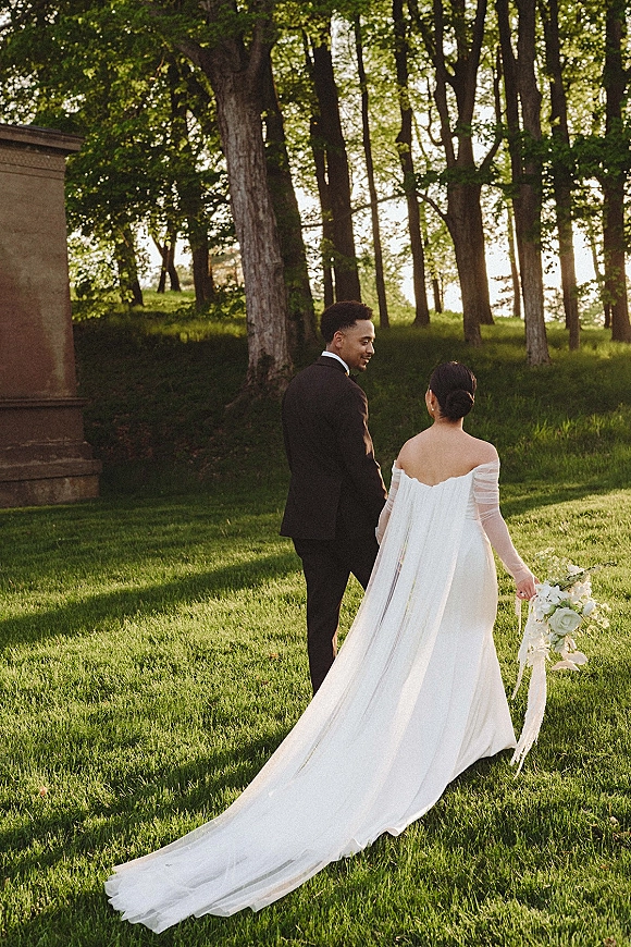 Couple portrait of bride and groom walking hand in hand, her long train and cascading bouquet with ribbon flowing on a sunny lawn