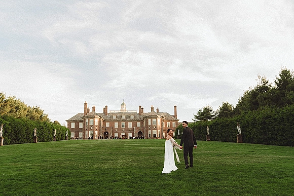 Couple portrait of bride and groom walking away holding hands, her wedding dress train flowing on a manor lawn with garden statues