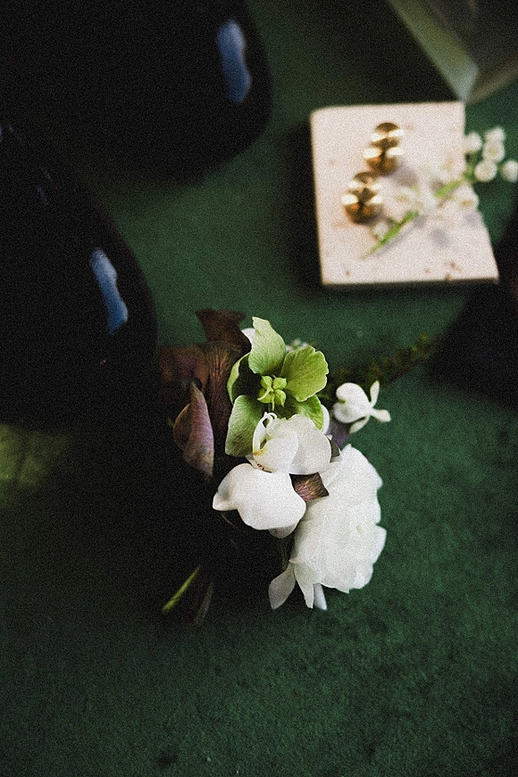 Wedding boutonniere with white flowers and green blooms pinned beside gold cufflinks on a shirt sleeve against a green fabric surface
