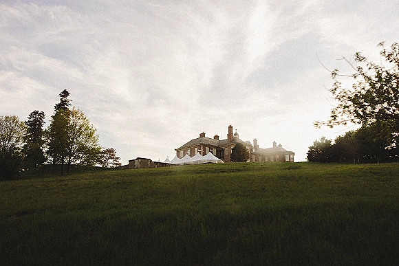 Wedding tent setup with a white event tent beside a stone wall, set on a sunny estate lawn with manor, hillside, and trees behind