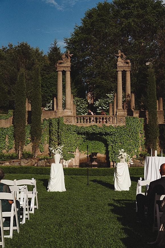 Outdoor ceremony setup with garden wedding ceremony aisle runner, white folding chairs and florals on pedestals before an ivy stone wall