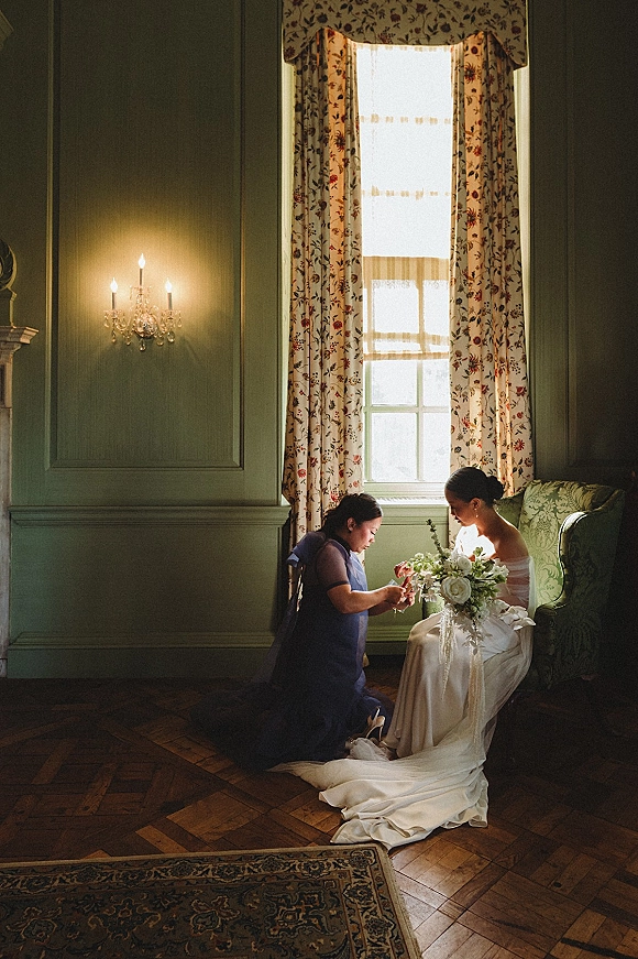 Bride getting ready with bridesmaid helping, seated in a strapless satin dress holding a white rose bouquet by a tall window in a classic room