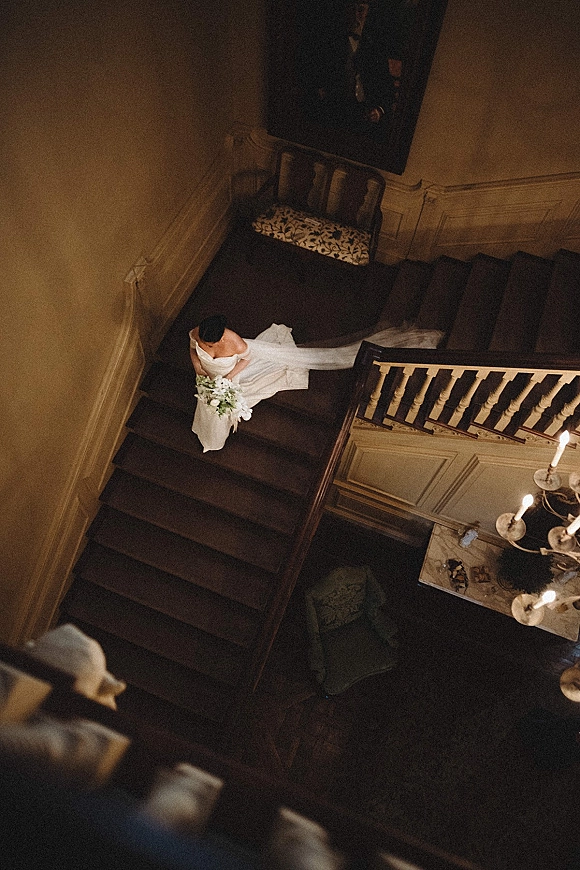 Bridal portrait of a bride on staircase with an off shoulder gown and long train, holding a white and green bouquet amid lit candlesticks.