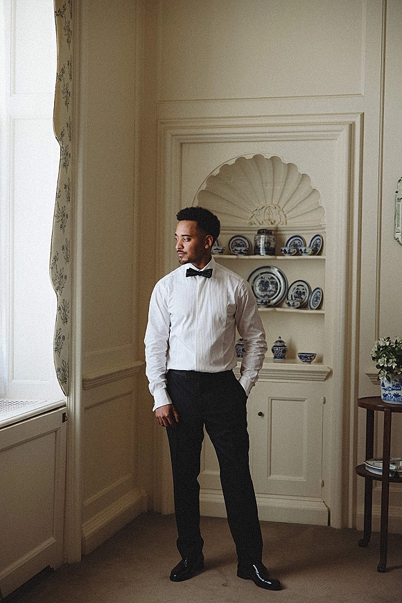 Groom portrait in a bow tie, hands in pockets, standing by window curtains with soft light and china display shelves behind
