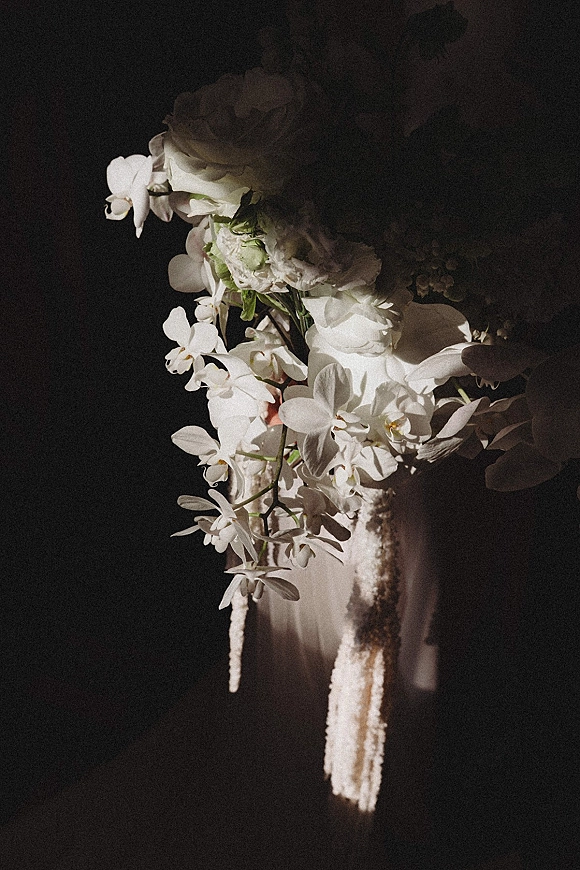 Wedding bouquet of white orchids and roses with trailing amaranthus accent cascading against a dark, shadowed backdrop
