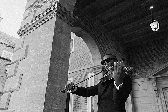 Wedding musician in a tuxedo and sunglasses playing violin with bow under a stone archway in a courtyard with hanging lanterns