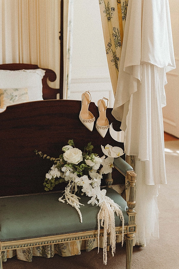 Wedding details with bridal shoes and bouquet on an upholstered bench, hanging dress beside a bed in soft window light