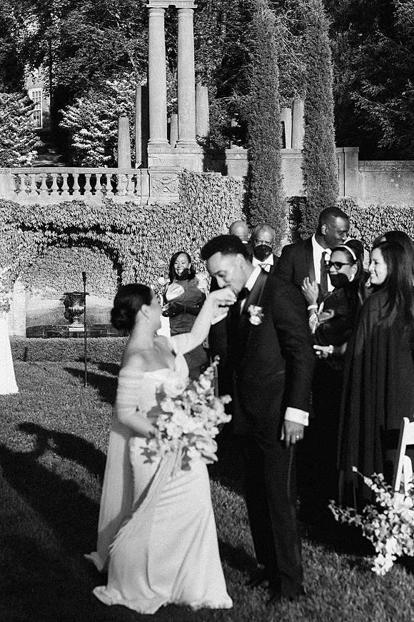 Wedding recessional as bride and groom exit, she holds a bouquet while masked guests cheer along a sunlit garden aisle with hedges
