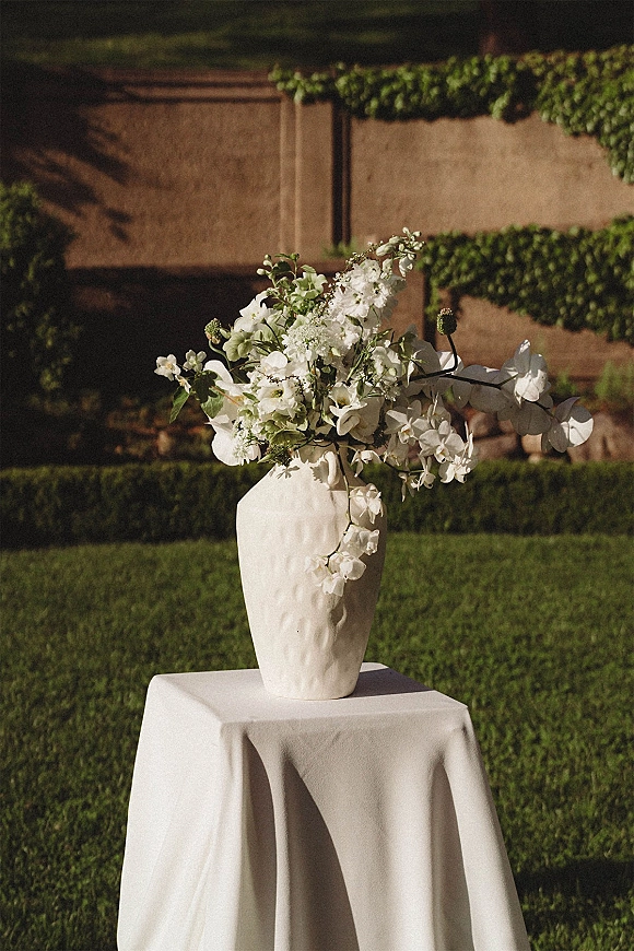 Wedding floral arrangement in a ceramic vase centerpiece with white blooms, greenery, and orchid stems on a pedestal table outdoors