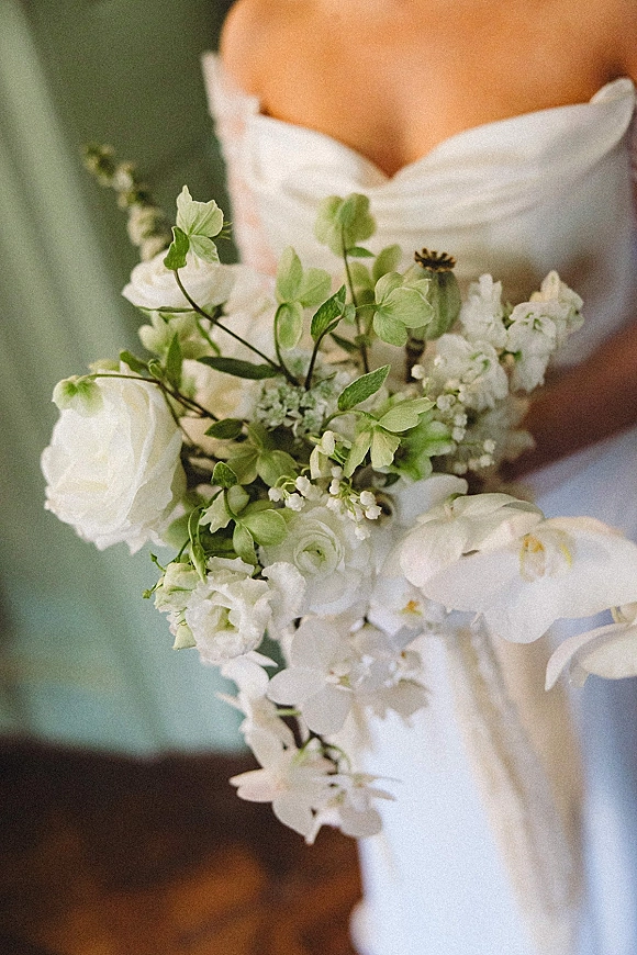 Bridal bouquet of white orchids and roses with ranunculus and greenery held against a strapless wedding dress in a soft indoor setting