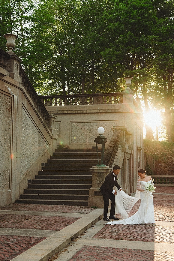Couple portrait of bride and groom portrait on a sunlit stone staircase, bride holding a white bouquet as groom lifts her dress train