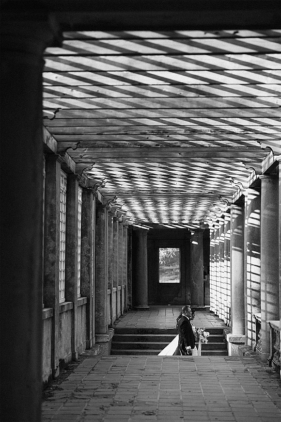Couple portrait in black and white wedding portrait style, bride in gown and veil holding bouquet beside groom in suit under pergola shadows