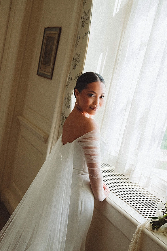 Bridal portrait of a bride by window in an off-the-shoulder dress with veil, sheer gloves, pearl earrings, holding a bouquet by curtains