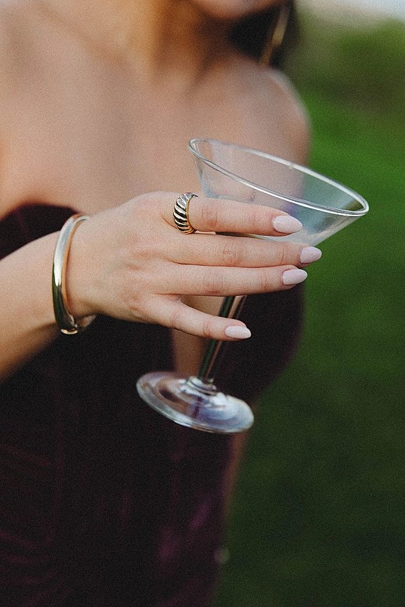Cocktail close-up in a martini glass wedding moment, held by a guest with manicured nails, gold ring and bangle, greenery blurred behind