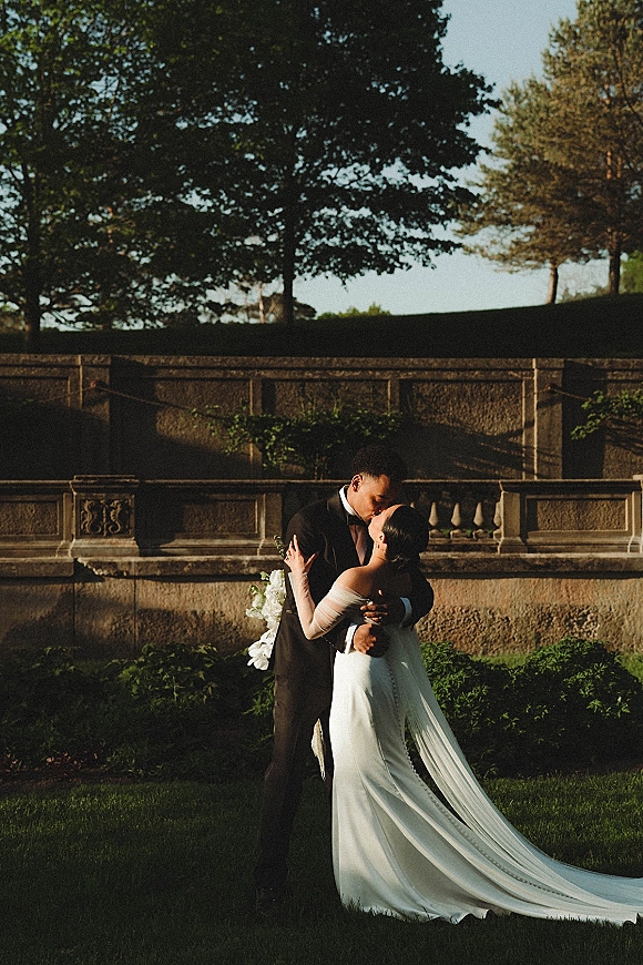 Wedding kiss portrait of bride and groom kissing on a stone terrace wall, her long train and white bouquet against a garden lawn at golden hour
