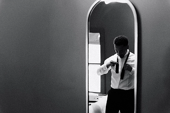Groom getting ready, tying a black tie in the mirror with cufflinks on a crisp dress shirt, lit by soft bedroom window light