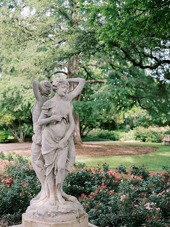 Garden statue in a classical garden sculpture setting with string lights overhead, surrounded by rose bushes and lush trees along a mulch path