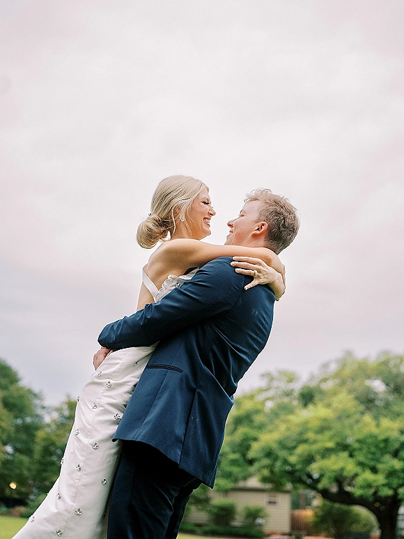 Couple portrait of bride and groom embrace as he lifts her, bride laughing in a strapless embellished gown in an overcast park setting