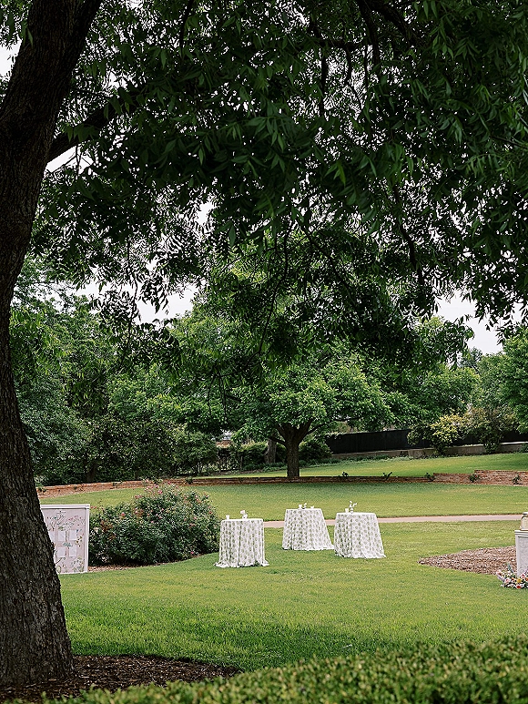 Cocktail hour setup with outdoor cocktail hour high-top tables, patterned linens, bud vases and place cards on a garden lawn under trees