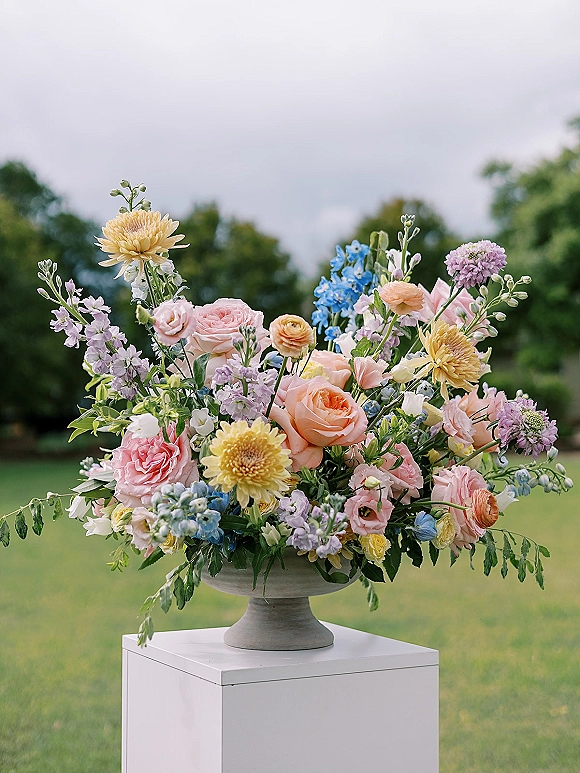 Wedding centerpiece of pastel roses, ranunculus and blue delphinium in a pedestal bowl on a white plinth, set on a lawn with trees under cloudy sky