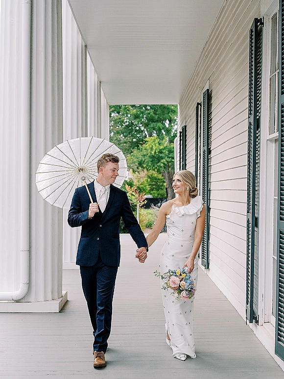 Couple portrait of bride and groom holding hands under a white parasol, she holds a pastel rose bouquet on a columned porch