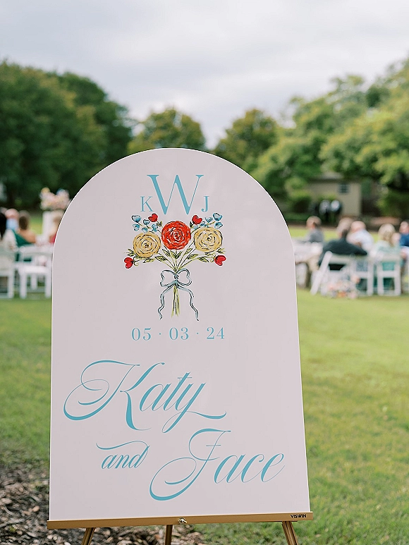 Wedding welcome sign with calligraphy lettering and monogram initials on an easel, with floral and ribbon accents on a grassy lawn by ceremony seating