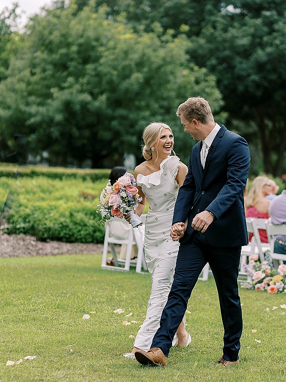Wedding recessional as bride and groom walking hand in hand, laughing down a petal-strewn garden aisle with seated guests and chairs