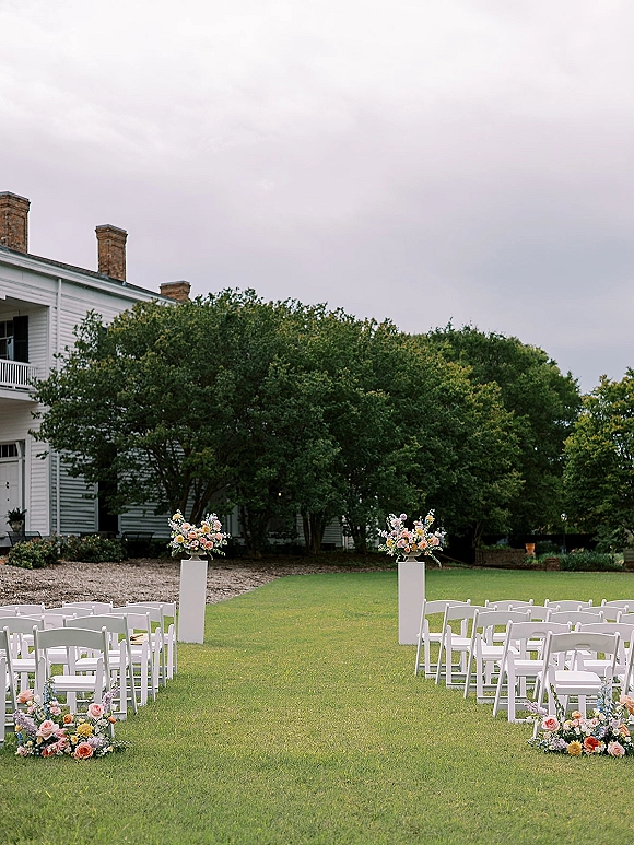 Ceremony setup with outdoor ceremony seating of white folding chairs and peach-pink aisle florals on a lawn, with trees and a white house behind