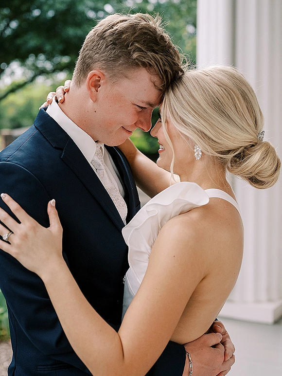 Couple portrait of bride and groom embrace with foreheads touching, groom in navy suit, bride in ruffle dress by greenery and white column