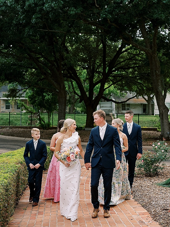 Wedding party portrait with bride and groom walking, bride holding bouquet, flanked by bridesmaids and groomsmen on a brick garden walkway