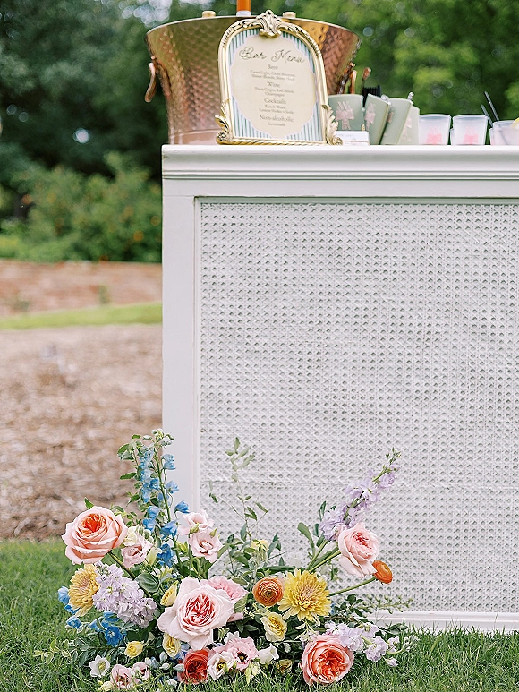 Wedding bar setup with a wedding bar menu sign, copper ice bucket, and floral arrangement on a white bar in an outdoor garden setting