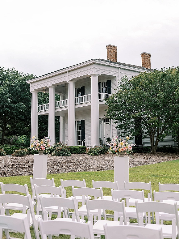 Outdoor ceremony setup with garden wedding ceremony seating, white folding chairs and floral pedestals on a lawn before a columned house