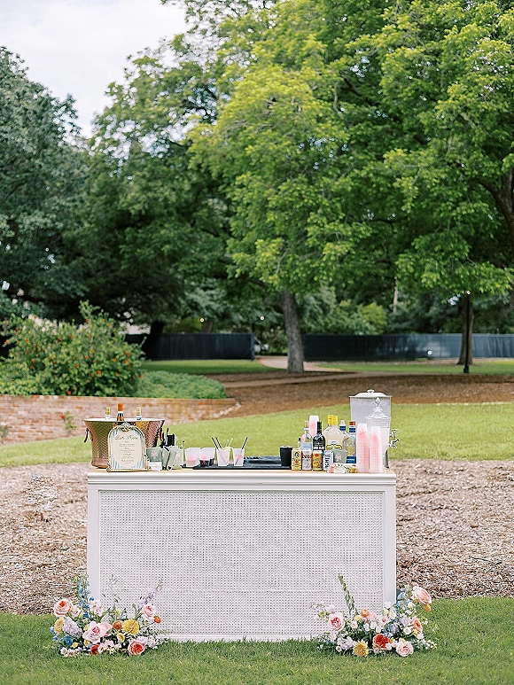 Wedding bar setup with liquor bottles, ice bucket, menu sign, and floral arrangements on a cart in a garden lawn by a brick wall