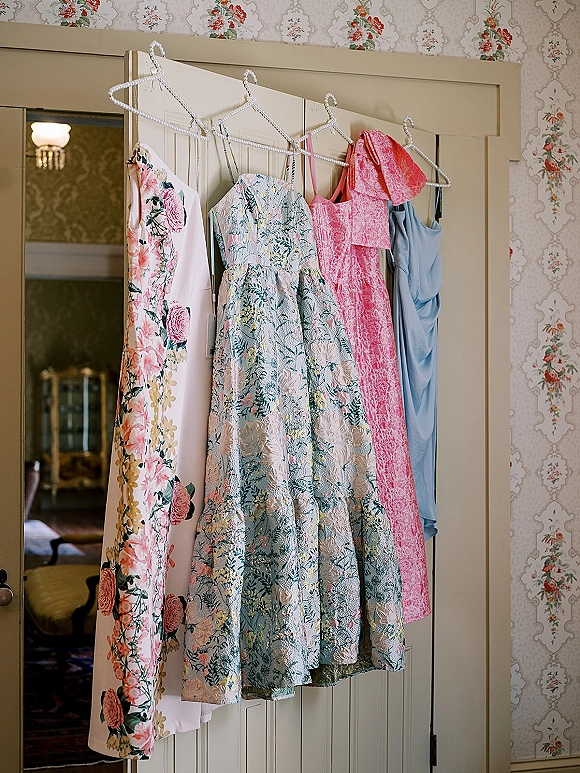 Bridesmaid dresses, mismatched bridesmaid dresses on beaded hangers with floral, jacquard, and satin styles against floral wallpaper by a door