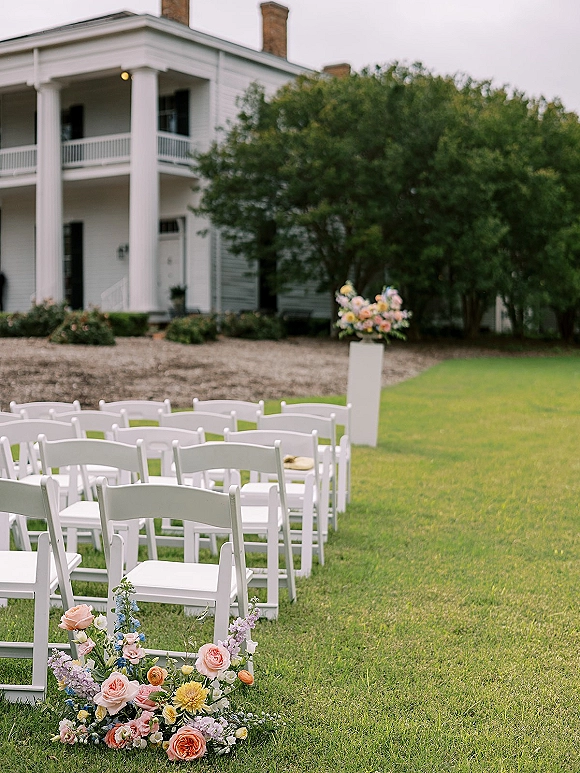 Outdoor ceremony setup with white wedding chairs in neat rows, framed by pedestal florals on a green lawn near a white house porch